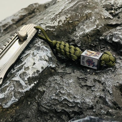 Rose Skull Titanium Lanyard Bead: bead on green cord beside a knife display prop (gallery view 02).