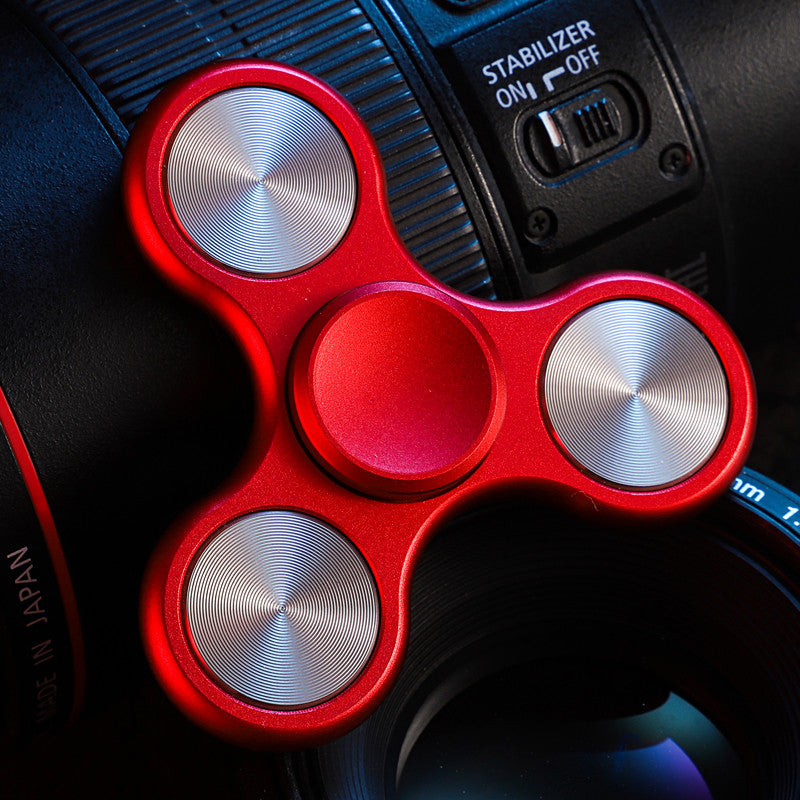 Red three-lobe spinner with silver-tone circular inlays; background camera is a photo prop.