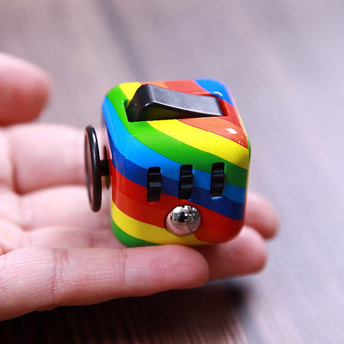 Rainbow-striped fidget cube held in one hand, showing its black rocker switch, three ridged rollers, metallic ball and round joystick.